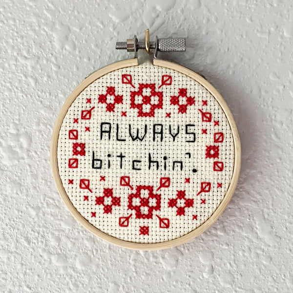 Hand embroidery of the words "always bitchin'" in black thread surrounded by red cross-stitch patterns. Art from Osseous Design by independent artist Makena Peet.