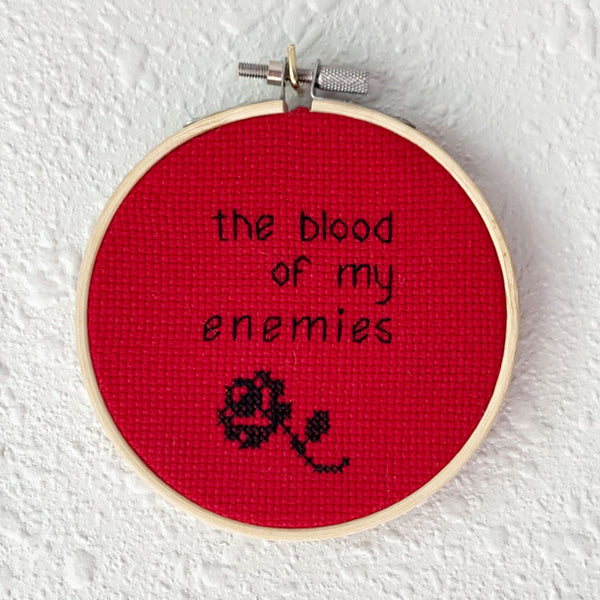 Hand embroidery of the words "the blood of my enemies" and a rose in black thread on red cross-stitch fabric. Art from Osseous Design by independent artist Makena Peet.