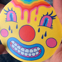 Close up image of a circular sticker of a clown-like emoji with a dripping rainbow forehead. Art from Osseous Design by independent artist Makena Peet.