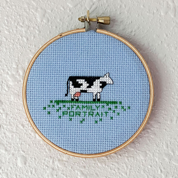 Hand embroidery of a cow over the words "family portrait" on light blue cross-stitch fabric. Art from Osseous Design by independent artist Makena Peet.