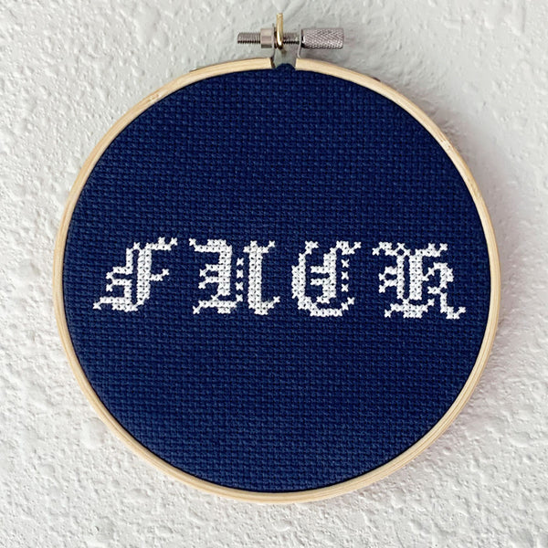 Hand embroidery of a popular curse word in white on dark blue cross-stitch fabric. Art from Osseous Design by independent artist Makena Peet.